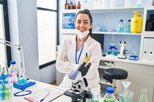Young beautiful hispanic woman scientist wearing medical mask sitting with arms crossed gesture at laboratory