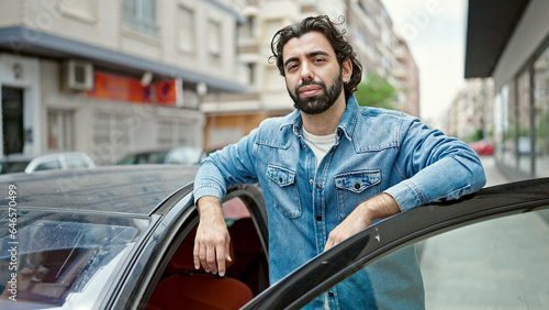 Young hispanic man leaning on car door with serious face at street