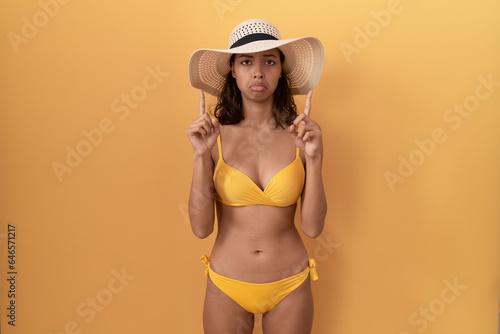 Young hispanic woman wearing bikini and summer hat pointing up looking sad and upset, indicating direction with fingers, unhappy and depressed.