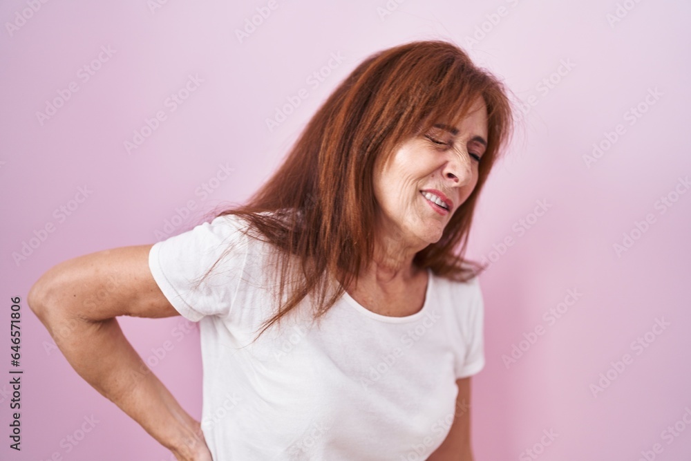 Middle age woman standing over pink background suffering of backache, touching back with hand, muscular pain Middle age woman standing over pink background suffering of backache, touching back with hand, muscular pain