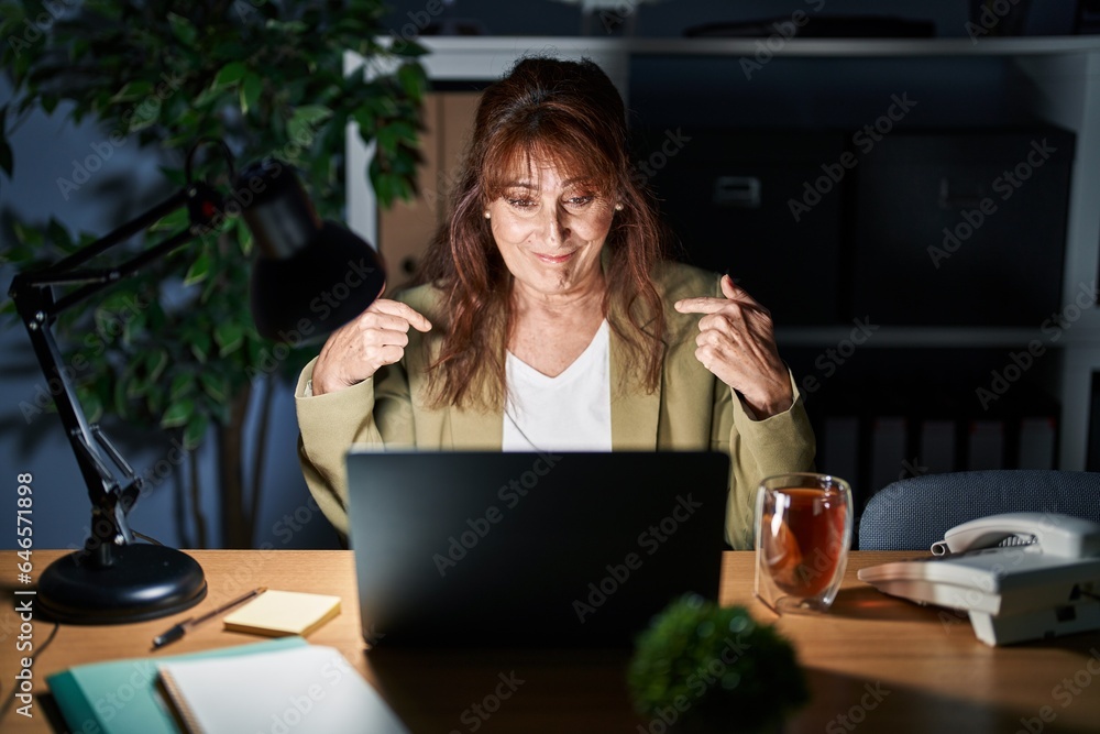Middle age hispanic woman working using computer laptop at night looking confident with smile on face, pointing oneself with fingers proud and happy.