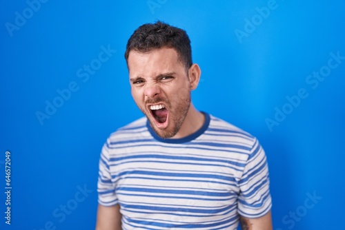 Young hispanic man standing over blue background angry and mad screaming frustrated and furious, shouting with anger. rage and aggressive concept.