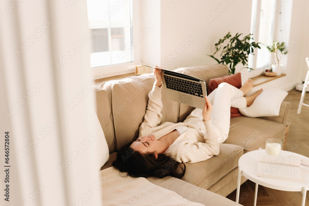 A young businesswoman in casual clothes works online from home using a laptop and wireless technology while sitting on a sofa in a cozy room at home. Selective focus A young businesswoman in casual clothes works online from home using a laptop and wireless technology while sitting on a sofa in a cozy room at home. Selective focus