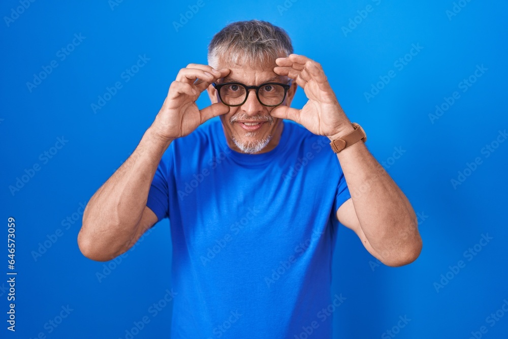 Hispanic man with grey hair standing over blue background trying to open eyes with fingers, sleepy and tired for morning fatigue Hispanic man with grey hair standing over blue background trying to open eyes with fingers, sleepy and tired for morning fatigue