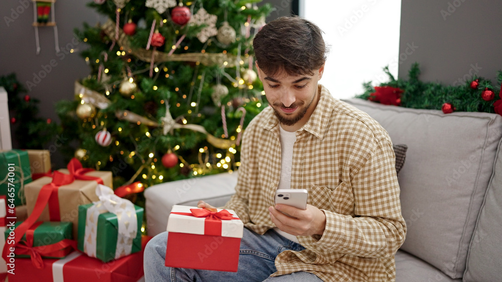 Young arab man holding gift using smartphone sitting by christmas tree at home Young arab man holding gift using smartphone sitting by christmas tree at home