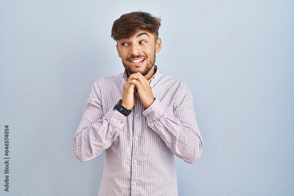 Arab man with beard standing over blue background laughing nervous and excited with hands on chin looking to the side Arab man with beard standing over blue background laughing nervous and excited with hands on chin looking to the side