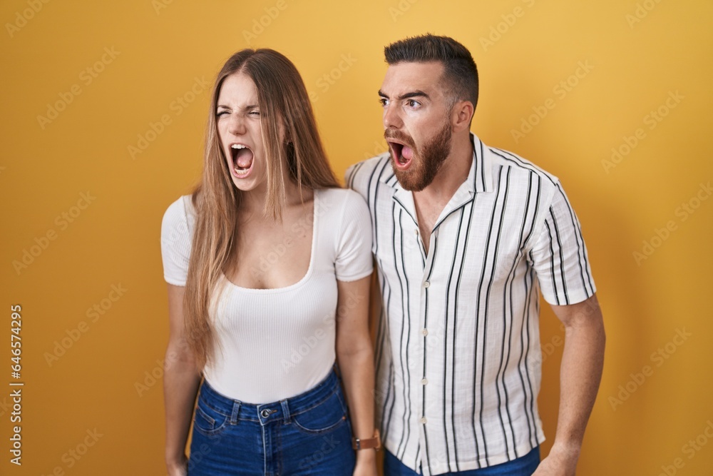 Young couple standing over yellow background angry and mad screaming frustrated and furious, shouting with anger. rage and aggressive concept. Young couple standing over yellow background angry and mad screaming frustrated and furious, shouting with anger. rage and aggressive concept.