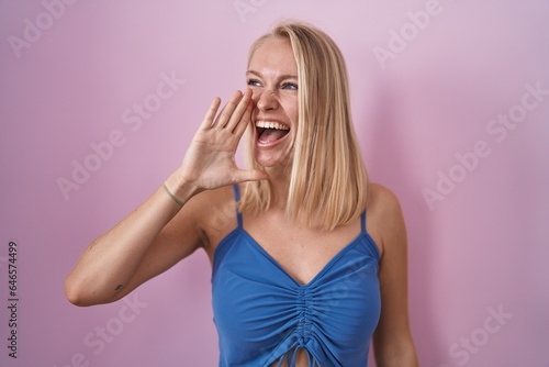 Young caucasian woman standing over pink background shouting and screaming loud to side with hand on mouth. communication concept.