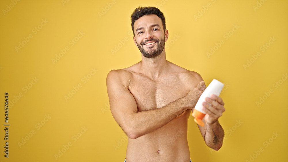 Young hispanic man tourist applying sunscreen shirtless over isolated yellow background Young hispanic man tourist applying sunscreen shirtless over isolated yellow background