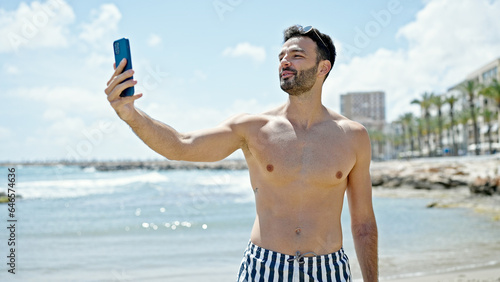 Young hispanic man tourist wearing swimsuit having video call at the beach