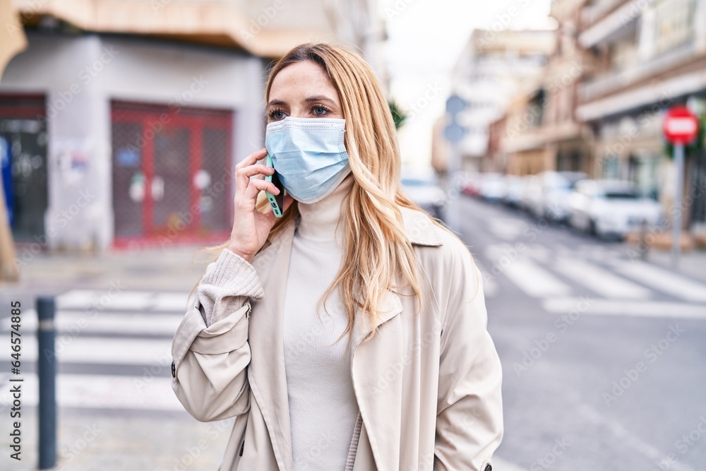 Obraz premium Young blonde woman wearing medical mask talking on smartphone at street