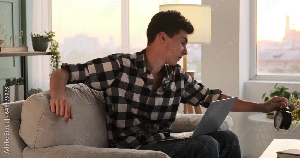 A young Caucasian man, dressed in casual attire, sits comfortably on a sofa with his laptop. Caught in a moment of tiredness, he yawns while balancing work and relaxation. A young Caucasian man, dressed in casual attire, sits comfortably on a sofa with his laptop. Caught in a moment of tiredness, he yawns while balancing work and relaxation.