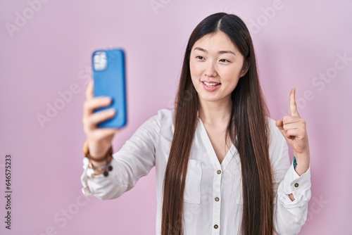 Chinese young woman taking a selfie photo with smartphone smiling with an idea or question pointing finger with happy face  number one