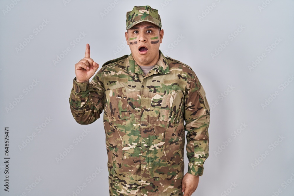 Hispanic young man wearing camouflage army uniform pointing finger up with successful idea. exited and happy. number one. Hispanic young man wearing camouflage army uniform pointing finger up with successful idea. exited and happy. number one.