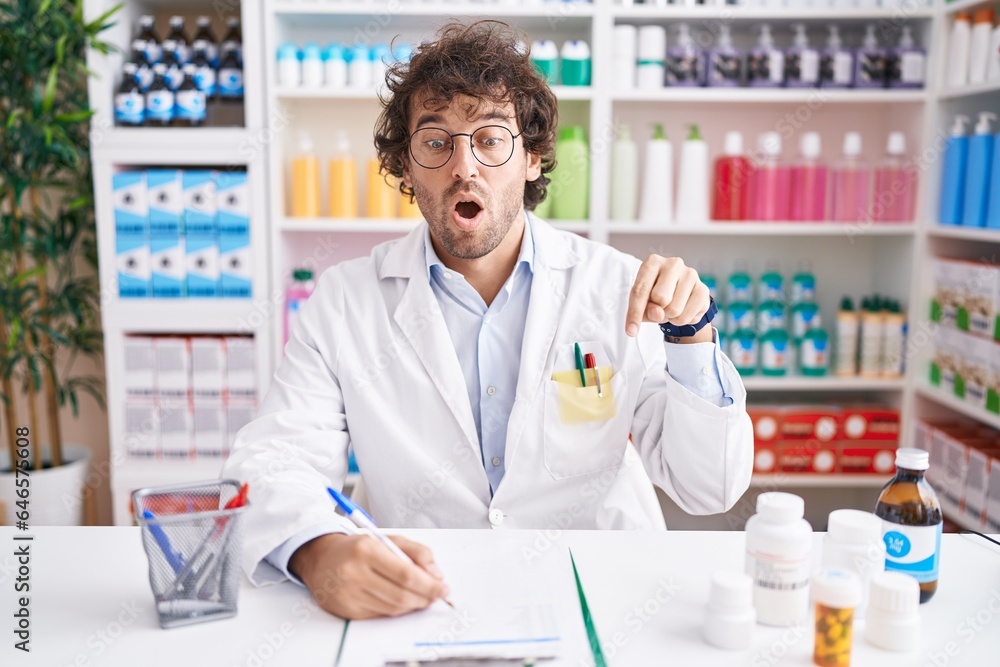 Hispanic young man working at pharmacy drugstore pointing down with fingers showing advertisement, surprised face and open mouth