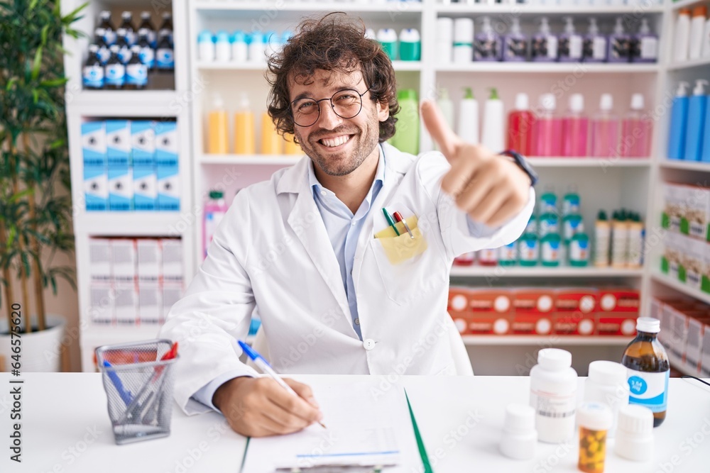 Hispanic young man working at pharmacy drugstore approving doing positive gesture with hand, thumbs up smiling and happy for success. winner gesture.