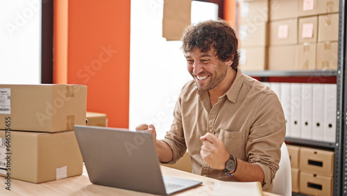 Young hispanic man ecommerce business worker typing on computer with winner gesture at office