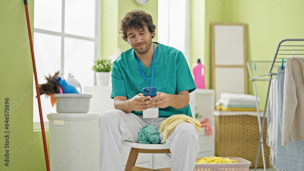 Young hispanic man professional cleaner using smartphone relaxing at laundry room
