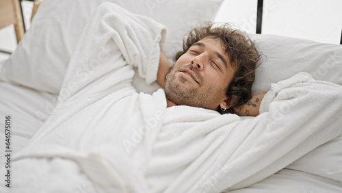 Young hispanic man wearing bathrobe relaxed on bed at bedroom