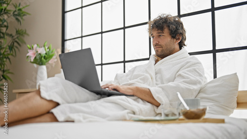 Young hispanic man wearing bathrobe using laptop at bedroom