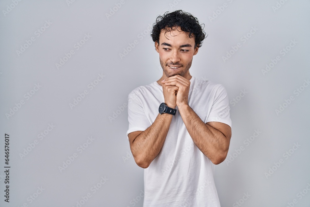 Hispanic man standing over isolated background laughing nervous and excited with hands on chin looking to the side Hispanic man standing over isolated background laughing nervous and excited with hands on chin looking to the side