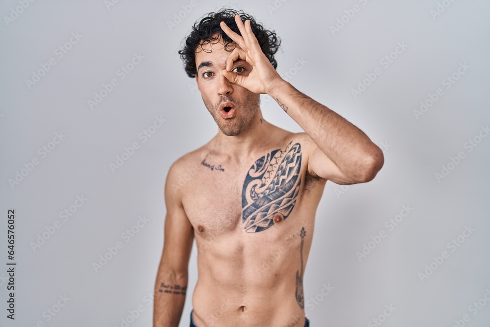 Fototapeta premium Hispanic man standing shirtless doing ok gesture shocked with surprised face, eye looking through fingers. unbelieving expression.