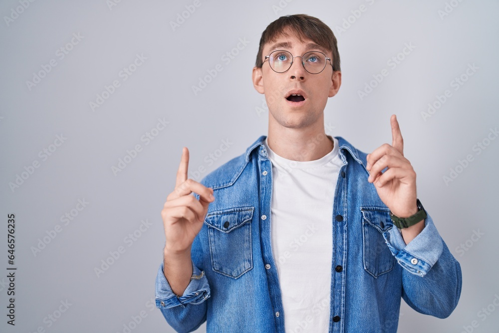 Caucasian blond man standing wearing glasses amazed and surprised looking up and pointing with fingers and raised arms. Caucasian blond man standing wearing glasses amazed and surprised looking up and pointing with fingers and raised arms.
