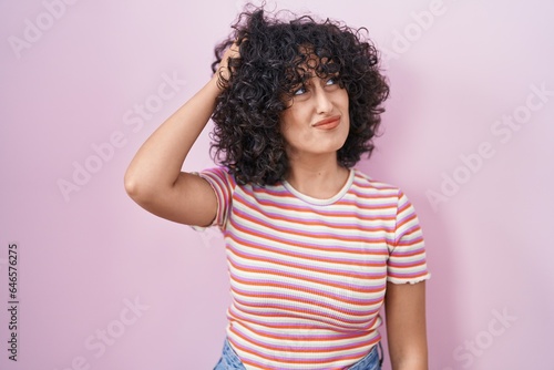 Young middle east woman standing over pink background confuse and wondering about question. uncertain with doubt, thinking with hand on head. pensive concept.
