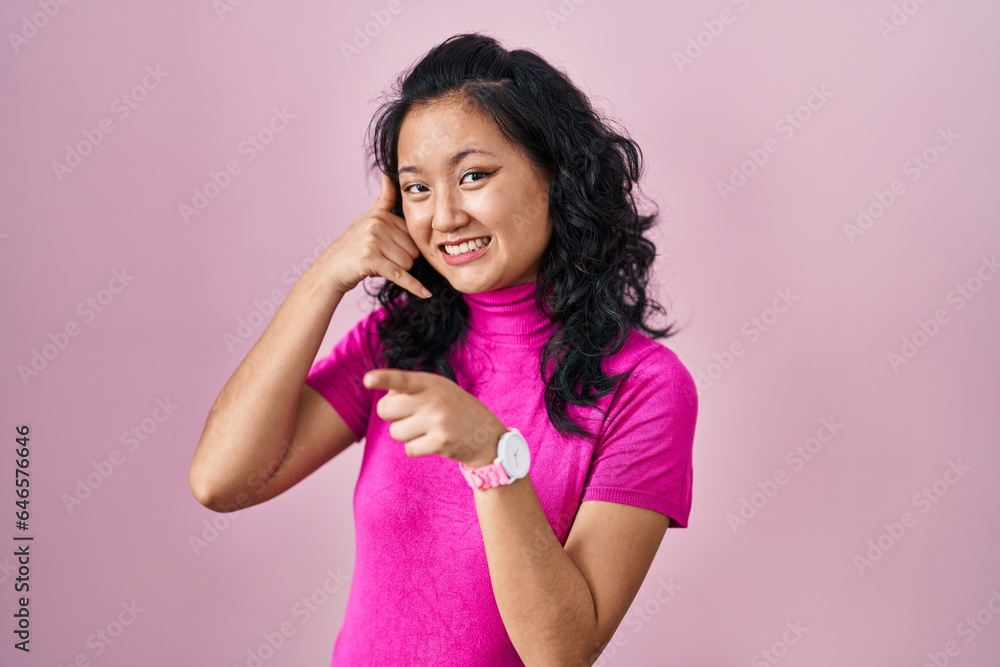Young asian woman standing over pink background smiling doing talking on the telephone gesture and pointing to you. call me. Young asian woman standing over pink background smiling doing talking on the telephone gesture and pointing to you. call me.