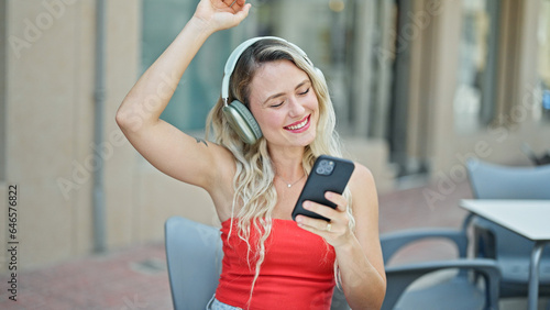 Young blonde woman listening to music and dancing sitting on table at coffee shop terrace