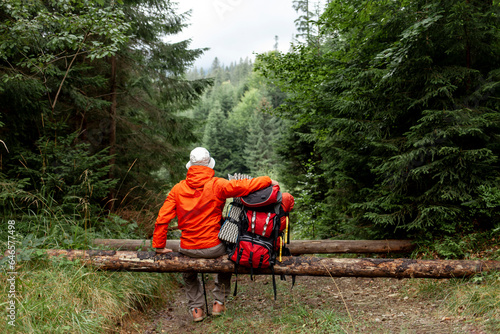 male tourist with backpack and hiking equipment sits resting in the forest, guy in jacket on mountain hike takes break