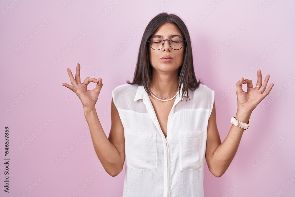 Brunette young woman standing over pink background wearing glasses relaxed and smiling with eyes closed doing meditation gesture with fingers. yoga concept. Brunette young woman standing over pink background wearing glasses relaxed and smiling with eyes closed doing meditation gesture with fingers. yoga concept.