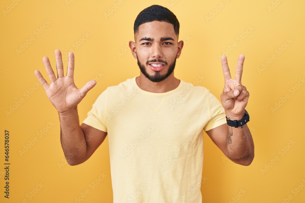 Young hispanic man standing over yellow background showing and pointing up with fingers number seven while smiling confident and happy. Young hispanic man standing over yellow background showing and pointing up with fingers number seven while smiling confident and happy.
