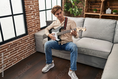 Young caucasian man playing electrical guitar sitting on sofa at home