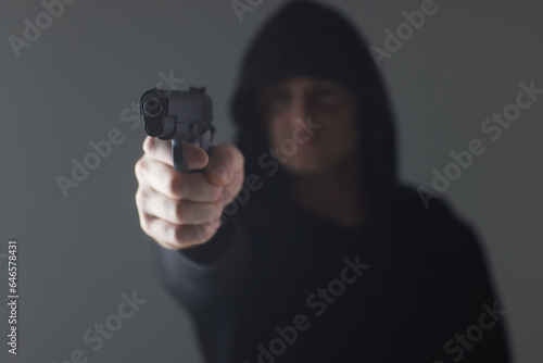 robber with gun aiming into camera. Man in hood threatens with firearm. Weapon in person's hands. Murderer or armed thief. Criminal with pistol.