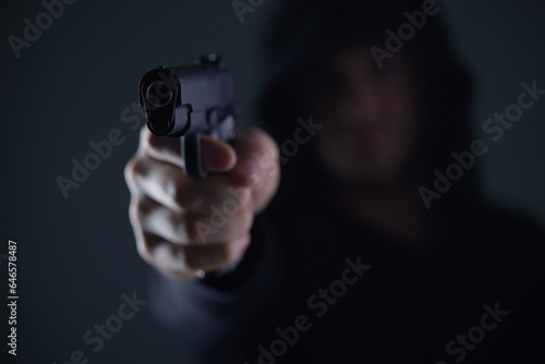 robber with gun aiming into camera. Man in hood threatens with firearm. Weapon in person's hands. Murderer or armed thief. Criminal with pistol.