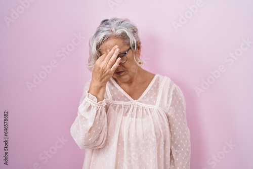 Middle age woman with grey hair standing over pink background tired rubbing nose and eyes feeling fatigue and headache. stress and frustration concept.