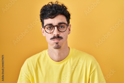 Hispanic man wearing glasses standing over yellow background puffing cheeks with funny face. mouth inflated with air, crazy expression.