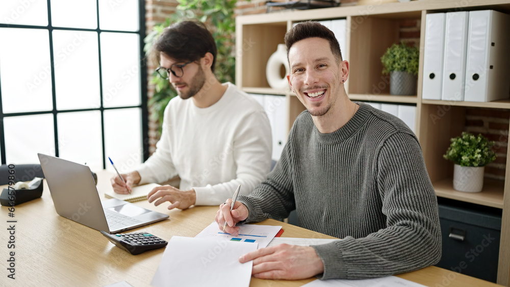 Two men business partners using laptop writing on document working at office Two men business partners using laptop writing on document working at office