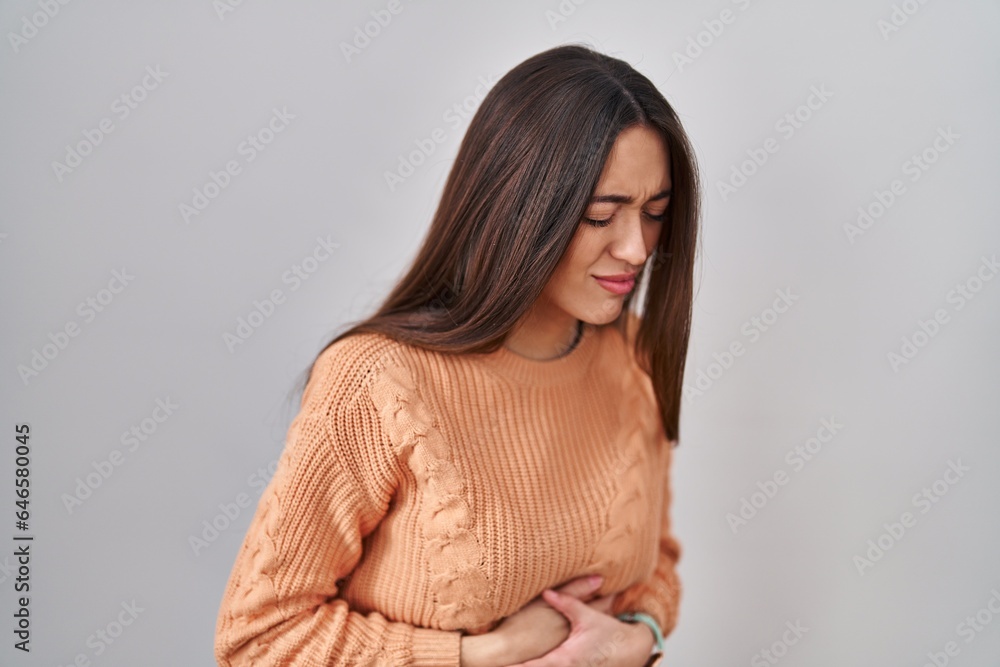 Obraz premium Young brunette woman standing over white background with hand on stomach because indigestion, painful illness feeling unwell. ache concept.
