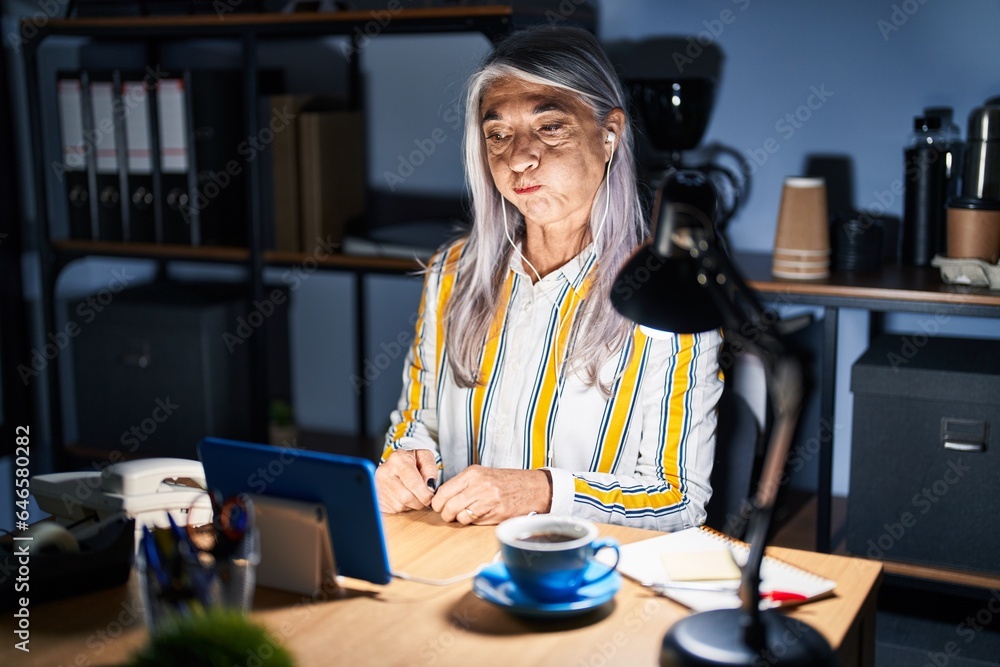Middle age woman with grey hair working at the office at night puffing cheeks with funny face. mouth inflated with air, crazy expression. - obrazy, fototapety, plakaty 