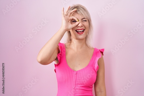 Young caucasian woman standing over pink background doing ok gesture with hand smiling, eye looking through fingers with happy face.