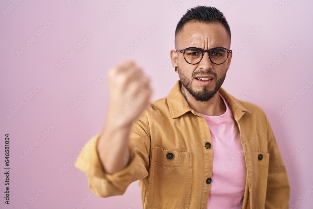 Young hispanic man standing over pink background angry and mad raising fist frustrated and furious while shouting with anger. rage and aggressive concept. Young hispanic man standing over pink background angry and mad raising fist frustrated and furious while shouting with anger. rage and aggressive concept.