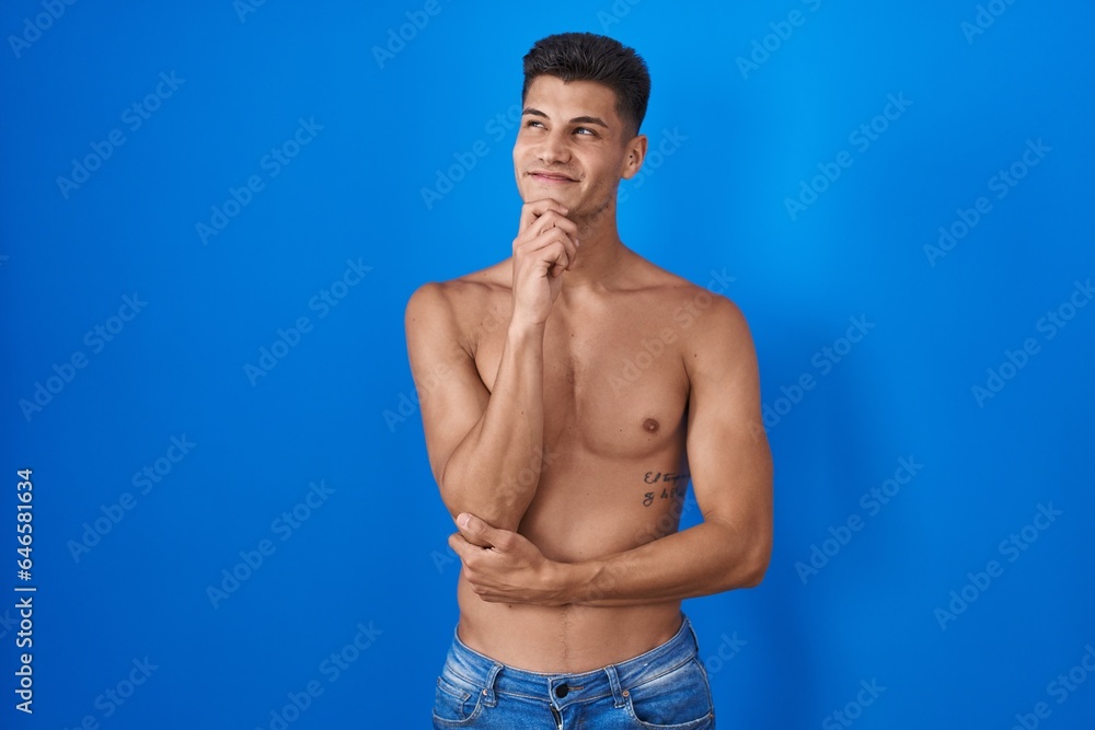 Young hispanic man standing shirtless over blue background with hand on chin thinking about question, pensive expression. smiling and thoughtful face. doubt concept.