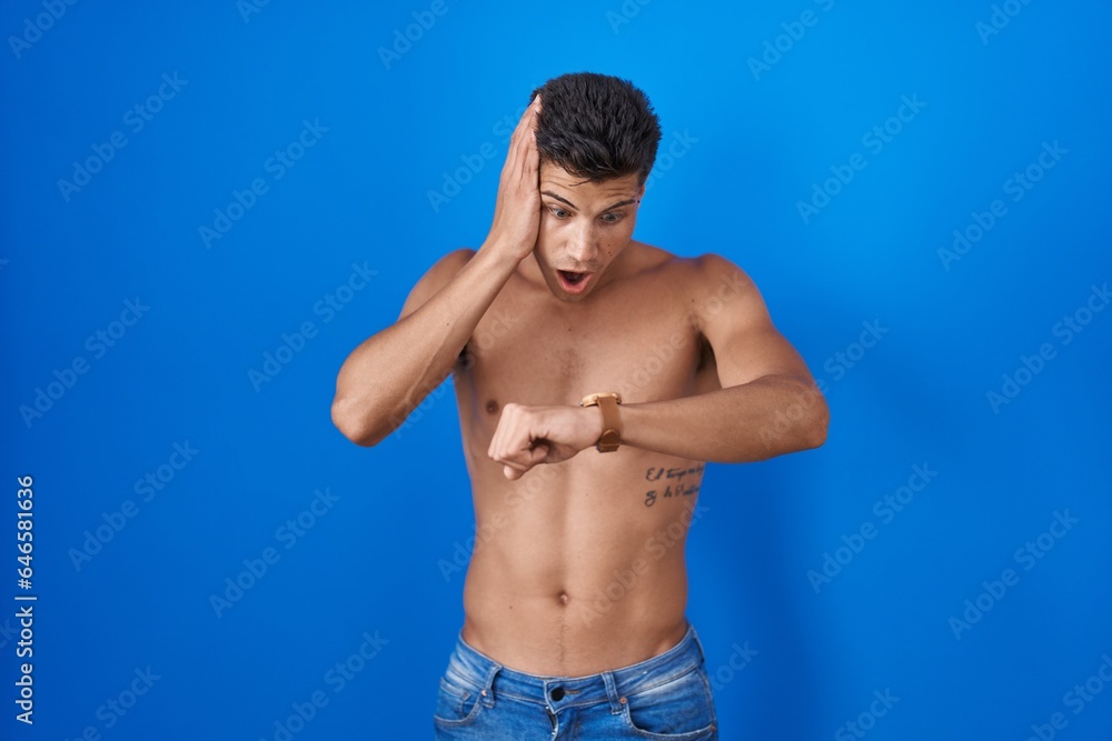 Young hispanic man standing shirtless over blue background looking at the watch time worried, afraid of getting late