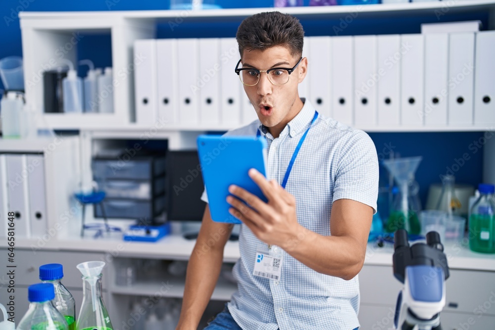 Young hispanic man working at scientist laboratory doing video call scared and amazed with open mouth for surprise, disbelief face