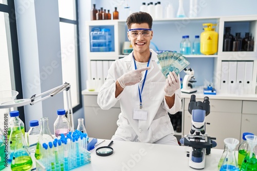 Young hispanic man working at scientist laboratory holding brazilian reals smiling happy pointing with hand and finger
