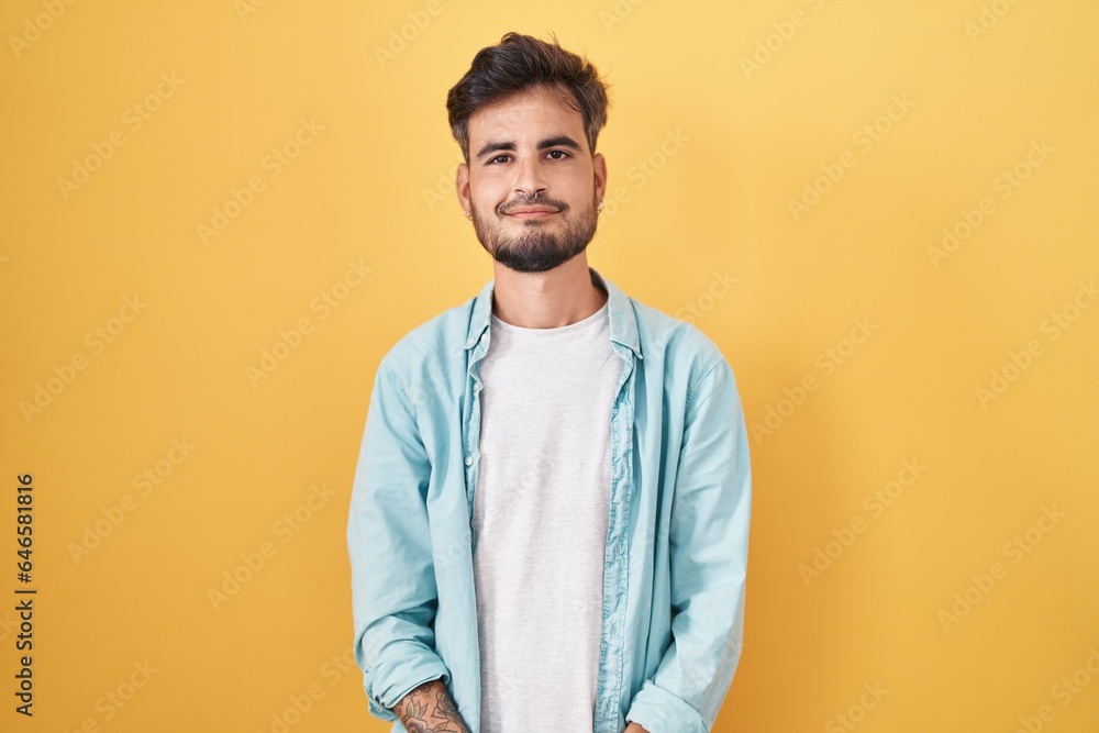 Young hispanic man with tattoos standing over yellow background with hands together and crossed fingers smiling relaxed and cheerful. success and optimistic Young hispanic man with tattoos standing over yellow background with hands together and crossed fingers smiling relaxed and cheerful. success and optimistic