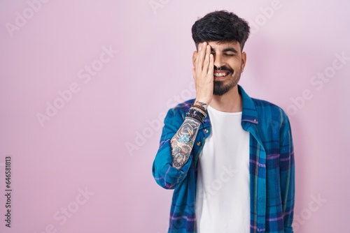 Fototapeta Naklejka Na Ścianę i Meble -  Young hispanic man with beard standing over pink background covering one eye with hand, confident smile on face and surprise emotion.
