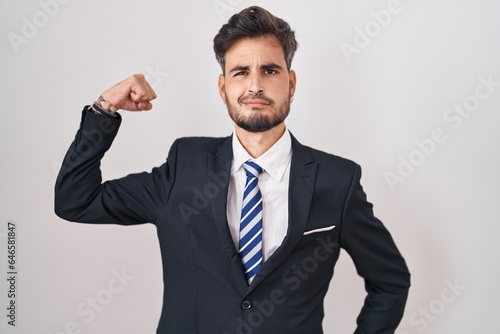 Young hispanic man with tattoos wearing business suit and tie strong person showing arm muscle, confident and proud of power